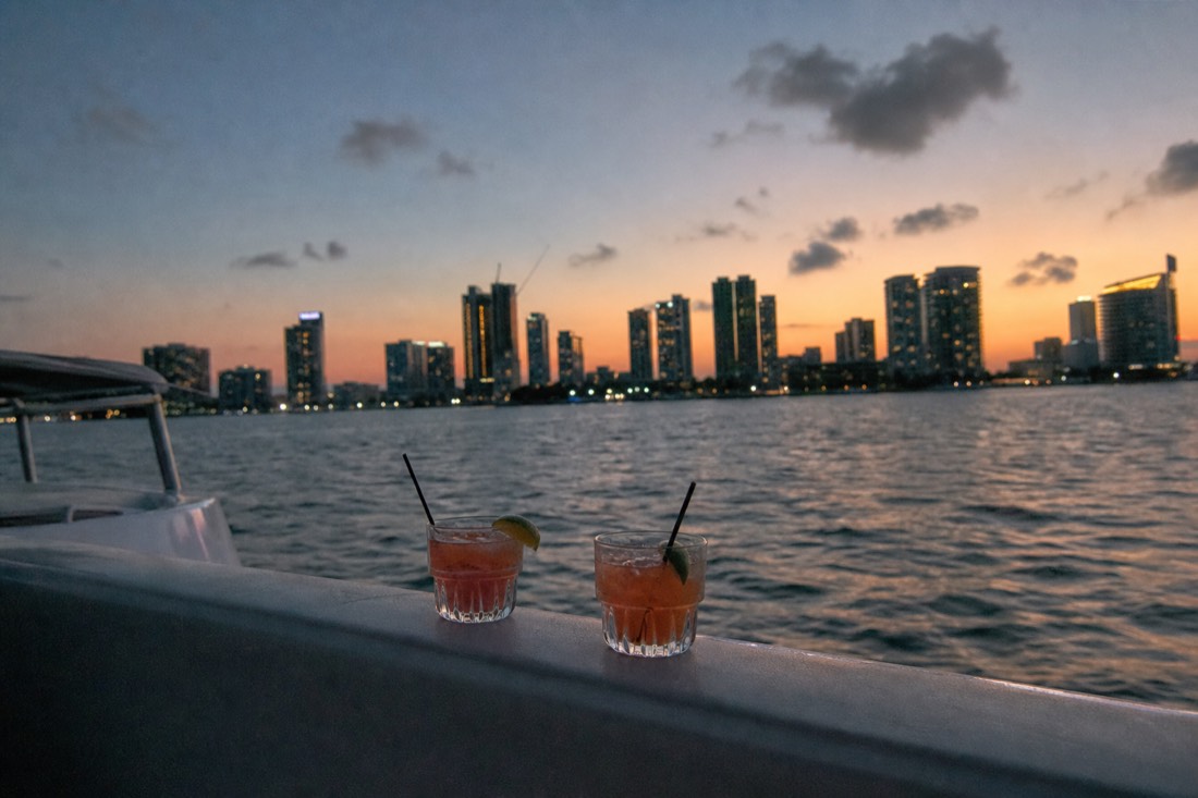 Cocktail glasses on a boat rail with the Miami Beach skyline and open water behind them at sunset, no text, no logos, no people
