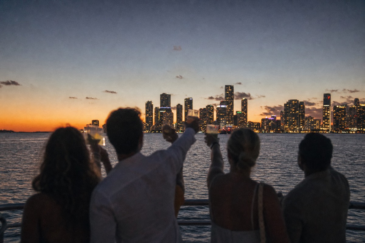 A group of friends toasting with drinks on an open boat deck as the Miami skyline lights up at dusk, no text, no logos, no people