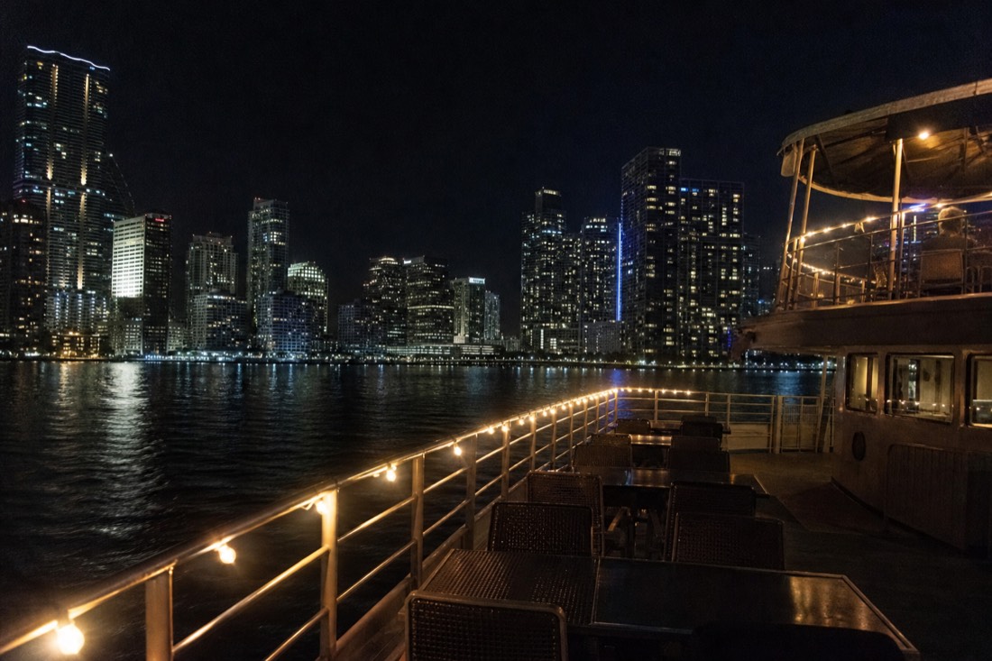 A double-deck passenger boat cruising past illuminated downtown Miami towers on Biscayne Bay at night, no text, no logos, no people