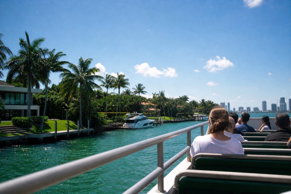 Passengers on the open upper deck of a sightseeing cruise boat passing celebrity homes along the Miami waterfront, no text, no logos, no people.