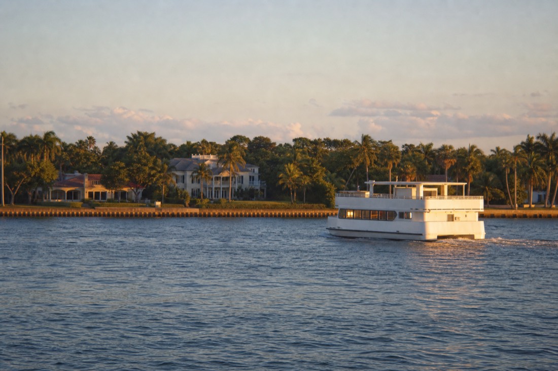 A double-deck passenger boat passing mansions on a tree-lined island in Biscayne Bay, no text, no logos, no people