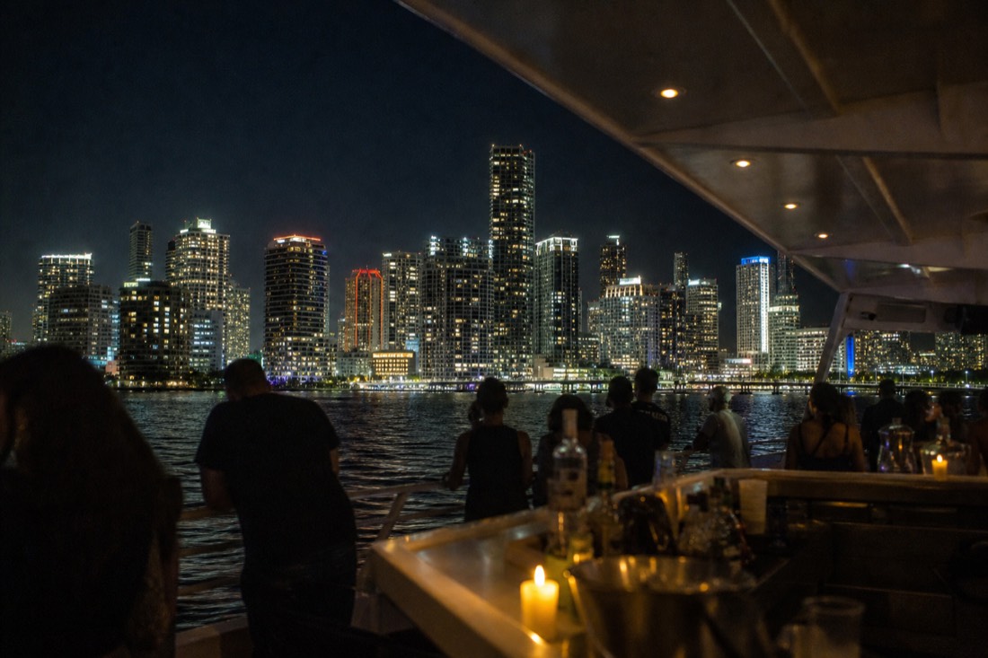 Open-deck cruise boat with an open bar setup crossing Biscayne Bay at night with the lit-up Miami skyline behind it