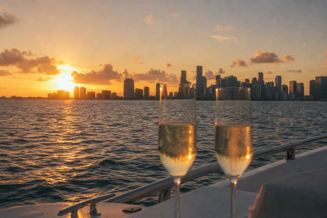 Two champagne glasses on a boat rail with Biscayne Bay and the Miami skyline glowing at sunset